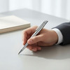 A silver metal pen held in a hand, resting on a grey surface with a book nearby. The pen has a sleek design and a logo.