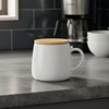 A white coffee mug with a wooden lid, placed on a countertop beside a coffee maker and bowls.