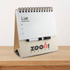 A tabletop notepad with a spiral binding, featuring a plain white notepad and a black marker. It has a logo on the base.