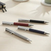 A collection of Geneva Metal Pens in grey, maroon, navy, and black with stainless steel accents, arranged on a desk.