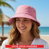 A pink bucket hat sits on a smiling woman's head with a beach backdrop and palm trees. The hat features a logo.