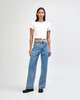 A woman stands wearing a white crop fitted T-shirt and blue jeans, with black shoes, against a light background.