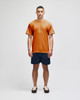 A man stands in a burnt orange short sleeve t-shirt paired with navy shorts, featuring a logo on the shirt.