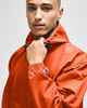 An orange packable anorak jacket worn by a model, featuring a hood and a branding logo on the sleeve.