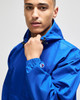 A blue packable anorak jacket worn by a model, featuring a hood and adjustable elements, with a logo on the sleeve.