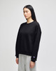 A black crewneck sweatshirt is displayed on a model, featuring a logo on the sleeve. The model wears grey pants.