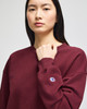 A burgundy women's crewneck sweatshirt featuring a logo on the sleeve. The model is standing and looking forward.