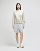 A model wearing a light grey crewneck sweatshirt and grey shorts, paired with white sneakers. The sweatshirt has a logo.
