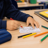 A hand holding a red crayon colours in a sketch on a small notepad, surrounded by coloured pencils and stationery.