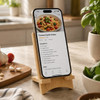 A wooden charging stand with a smartphone displaying a pasta recipe, surrounded by kitchen items and greenery.