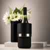 A bottle cooler in black with a silver band, featuring a wine bottle and a vase of flowers in the background.