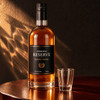 A bottle of premium blended whisky beside a clear shot glass, set on a wooden surface with textured lighting.