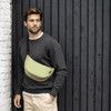 A waist bag in light green worn across a man's chest, with a casual grey sweater and beige pants, against a dark wooden background.