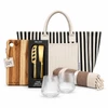 A striped tote bag with a wooden cheese board set, two glass tumblers, and a patterned blanket.