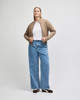 A woman wears a beige full-zip jacket over a white top, paired with wide-legged blue jeans. The jacket features a logo.