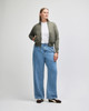 A woman wearing an olive green full-zip jacket over a white t-shirt, paired with light blue wide-leg jeans.