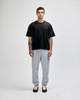 A man stands wearing a black heavyweight cotton unisex boxy t-shirt and grey sweatpants. The image features a logo on the t-shirt.