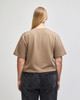A back view of a beige mid-length boxy top with short sleeves, paired with dark grey pants. The top features a logo.