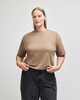 A mid-length boxy top in a light brown colour, featuring short sleeves and a round neckline. The model wears dark jeans.