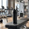 A drink bottle with a double-wall design in black, placed on a gym bench with exercise equipment in the background.