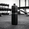 A drink bottle in black, standing upright on a gym floor with weights in the background.