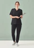 A woman wearing black women's Pulse jogger scrub pants and a matching top, holding a clipboard, standing against a light green background.