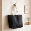 A black recycled beach tote bag with rope handles hangs on a hook against a textured wall.