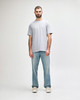 A man stands wearing a light grey Gildan Softstyle adult midweight T-shirt and blue jeans. The shirt has a logo.