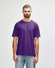 A man stands wearing a purple Gildan Softstyle adult T-shirt, paired with light blue jeans. The shirt has a logo.