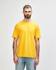 A person stands wearing a bright orange Gildan Softstyle adult T-shirt, paired with grey jeans. The shirt has a logo.