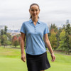 A woman is wearing a light blue polo shirt and a black skirt, standing on a golf course with trees in the background.