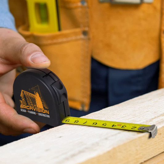 A black locking tape measure with a yellow measuring tape lies on a wooden surface, held by a person in work attire.