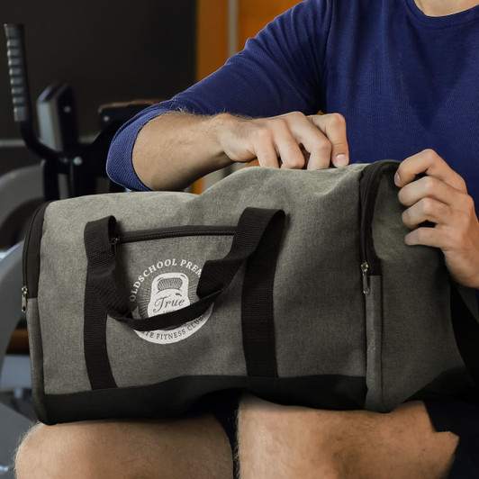 A grey duffle bag with handles and a logo, placed on a person's lap in a gym setting.