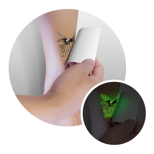 A hand applying a temporary glow-in-the-dark tattoo on a light-coloured arm. The design features a buzzing insect.