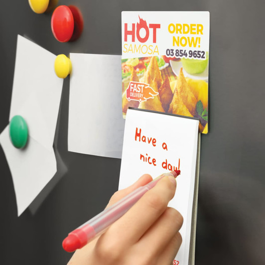 A full-colour A7 magnetic memo pad is attached to a fridge, with handwritten notes and colourful magnets around it.