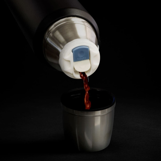 A drink bottle in black and silver, pouring a dark liquid into a stainless steel cup. It features a lid with a logo.