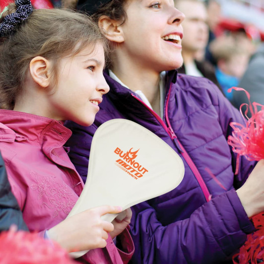 A foldable event fan in cream, held by a girl, with a logo displayed, surrounded by cheering spectators.