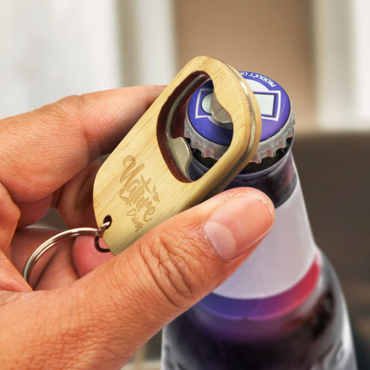 A drink bottle opener key ring made of wood, featuring a circular opening and a logo. The hand demonstrates its use.