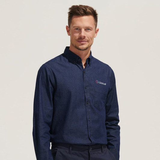 A man models a navy blue button-up denim shirt, featuring a logo on the chest pocket.