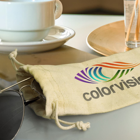 A cotton drawstring pouch in natural colour containing sunglasses, paired with a coffee cup and a wooden plate.