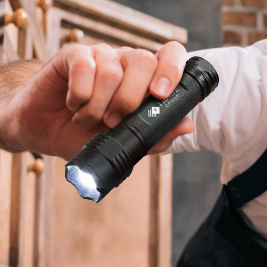 A black torch held in a hand, featuring a textured grip and a bright light at the front. It has a logo on its side.
