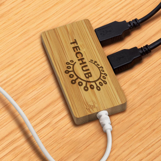 A bamboo USB hub with two black cables plugged in, placed on a wooden surface. The hub has a logo.