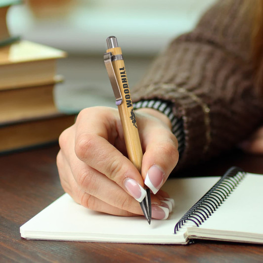Ancona Bamboo Inkless Pen with a wooden finish, held in a hand, writing in a notebook on a wooden table.