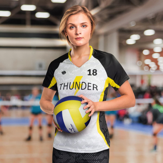 A woman in a black, white, and yellow volleyball jersey holds a volleyball. The jersey features a logo and number 18.