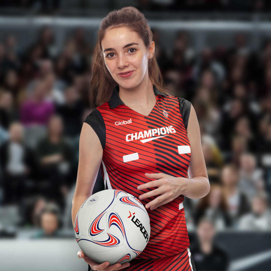A woman in a red and black netball top holds a ball, featuring a logo on the top. The background is blurry.