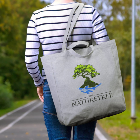 A grey tote bag featuring a tree design and a logo, carried by a person in a striped shirt on a pathway.