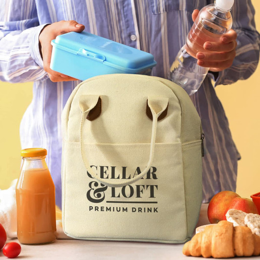 A yellow lunch bag with a logo, surrounded by a drink bottle, blue lunch box, juice, and apples.
