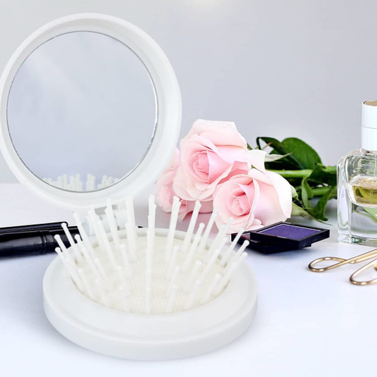 A compact white brush with a mirror, featuring bristles, placed among roses and beauty products.