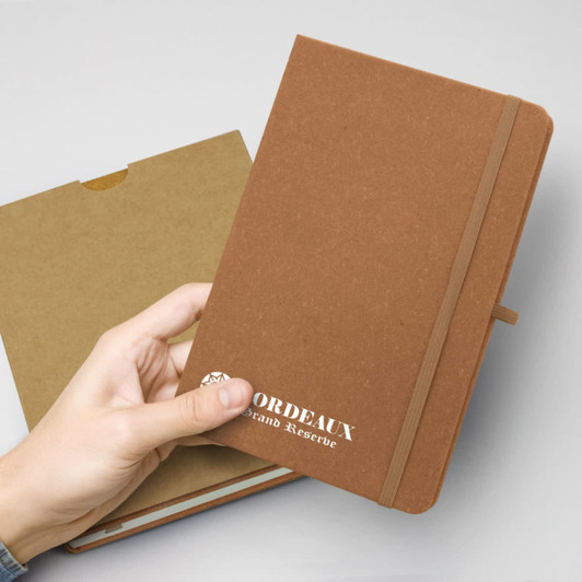 A hand holding a brown hardcover notebook with an elastic band, featuring a logo on the front.