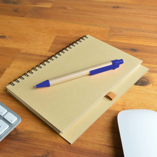 A brown spiral notebook with a blue pen resting on top, placed on a wooden surface alongside a keyboard and mouse.